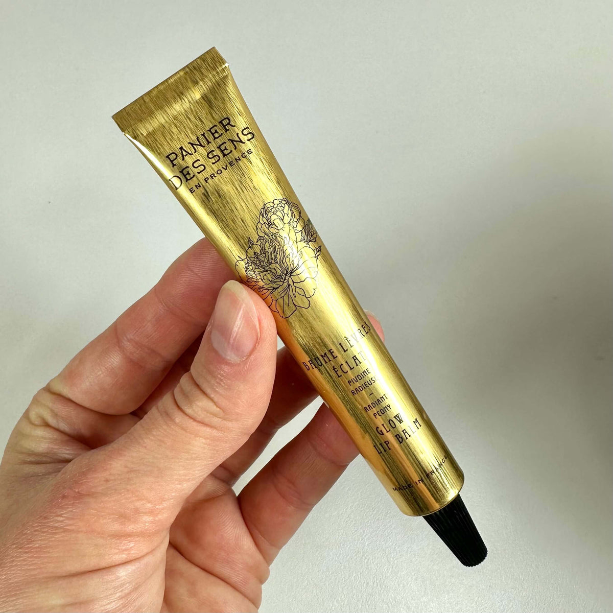 En hånd holder en gylden tube af Panier des Sens Glow Lip Balm mod en neutral baggrund. Tuben har et elegant design med blomsterillustration og sort tekst, der fremhæver produktets navn og egenskaber. Glow Lip Balm fugter læberne og giver en naturlig glød. Emballagen udstråler luksus og enkelhed, hvilket understreger produktets eksklusive og plejende karakter.