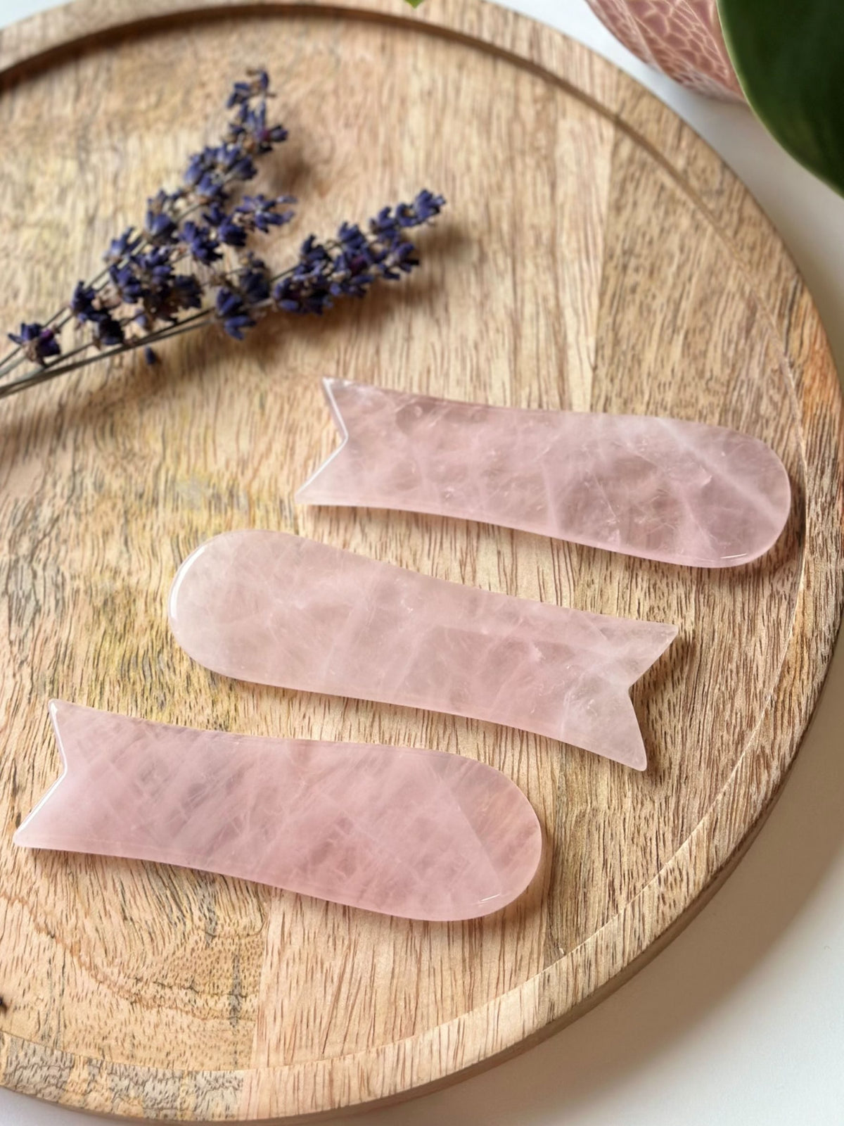Rosenkvarts Gua Sha værktøj på træbræt med lavendelblomst ved siden af.