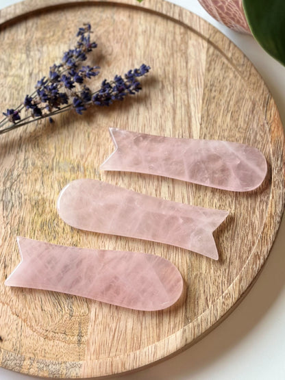 Rosenkvarts Gua Sha værktøj på træbræt med lavendelblomst ved siden af.