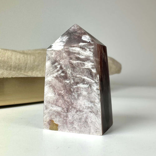 En poleret Lepidolit Mica-krystal med en pyramideformet top og varieret lilla farvenuance står på en lys overflade.