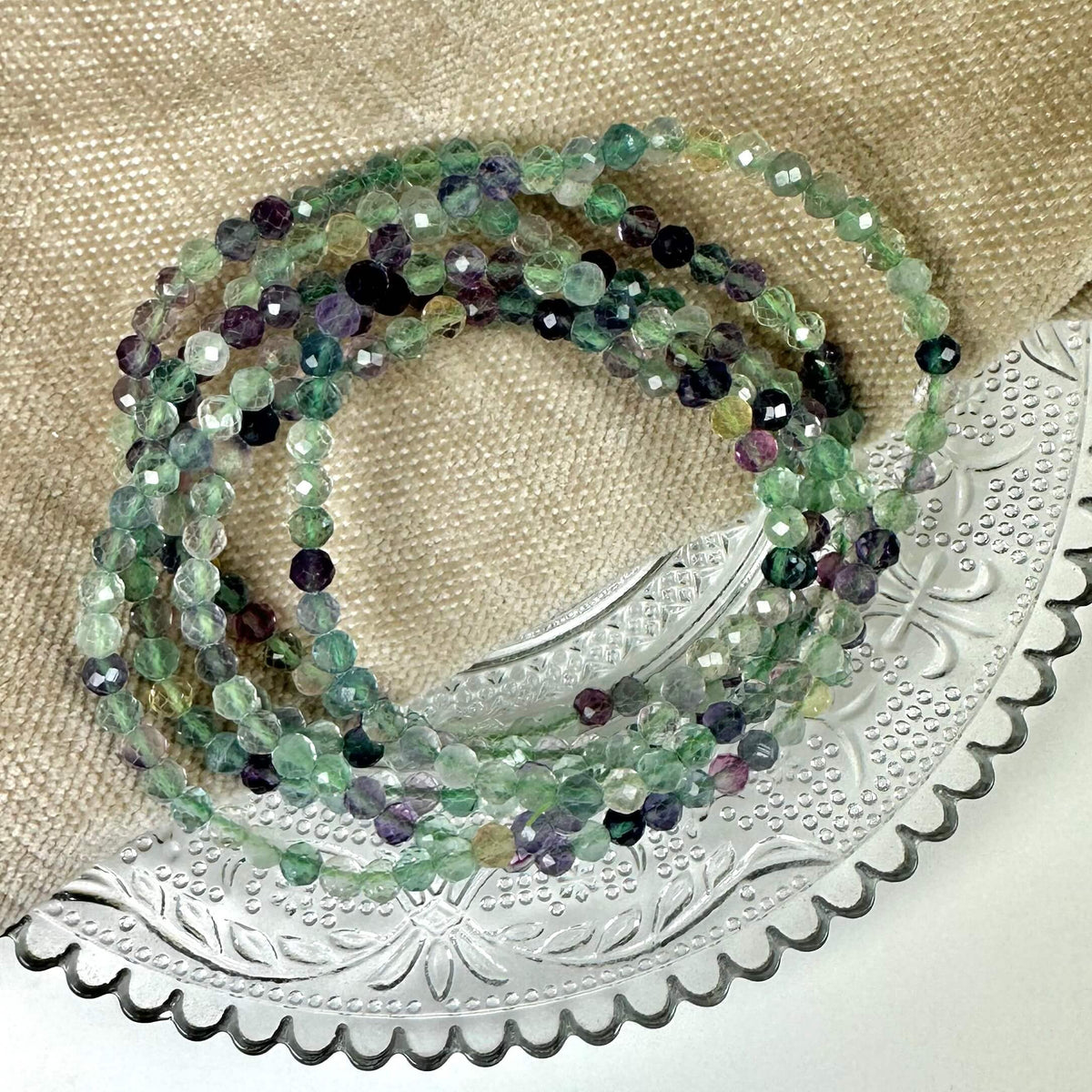 Fluorit armbånd med flerfarvede perler på en dekorativ bakke, placeret på en lys stofbaggrund.