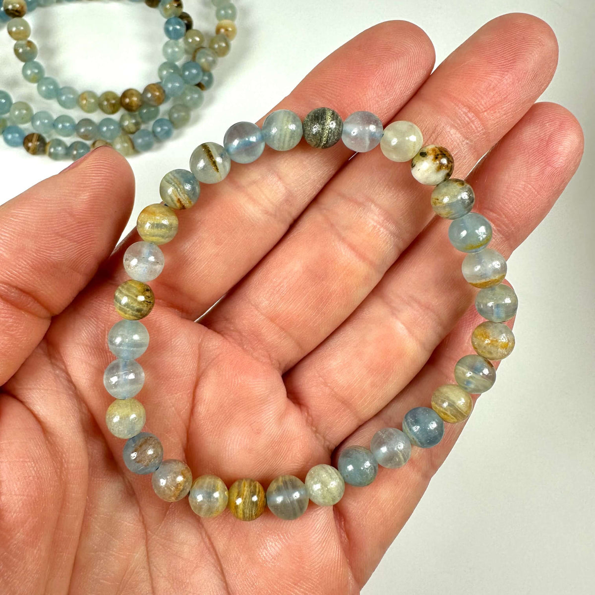 En hånd holder et Akvatin lemurie calcit armbånd 6mm med blålige og jordfarvede perler.