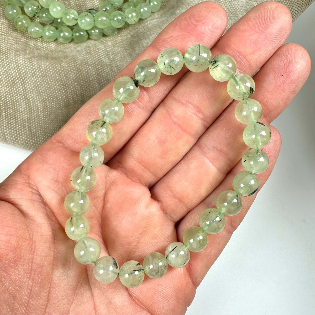 Prehnit armbånd 8mm perler i grønne nuancer vises i en hånd mod en beige baggrund.