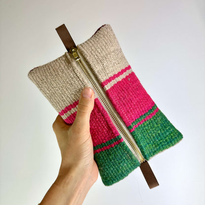 En hånd holder en farverig stofpung med pink, grønne og beige striber og en lynlås i midten.