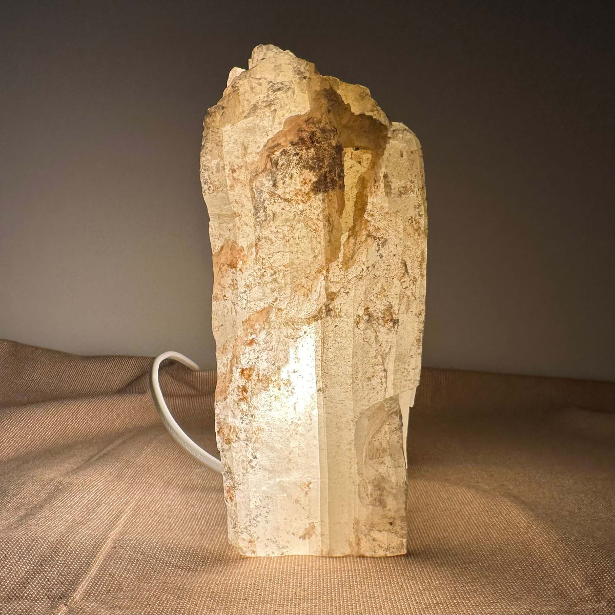 Mexicansk Selenit lampe nr. 38 står oprejst på en rustik tekstilbaggrund og udstråler et varmt, blødt lys gennem den transparente krystaloverflade. Lampens unikke, håndskårne form bibeholdes i en rå og organisk struktur med naturlige farvevariationer i hvid, beige og brun. Ledningen ses diskret i baggrunden. Et harmonisk designstykke, der kombinerer æstetik med spirituel og energirensende funktion.