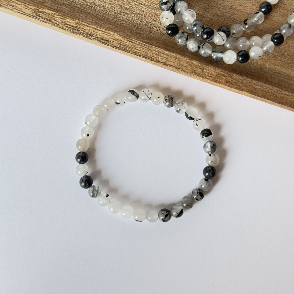 Turmalinkvarts armbånd med hvide og sorte perler placeret på hvid baggrund ved siden af træunderlag.