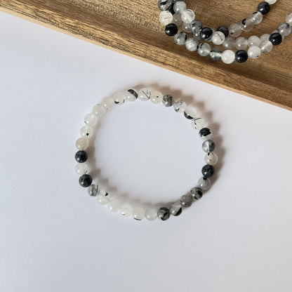 Turmalinkvarts armbånd med hvide og sorte perler placeret på hvid baggrund ved siden af træunderlag.