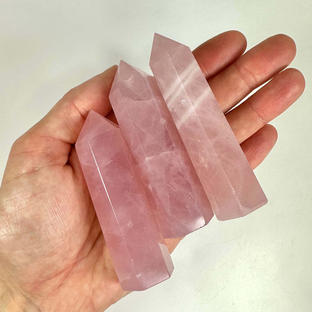 En hånd holder tre lyserøde rosenkvarts tårne med spidse ender, hver poleret til en glat finish. Krystallerne har en delikat rosa nuance og viser naturlige mønstre og gennemsigtighed. Rosenkvarts er kendt som kærlighedens sten og siges at fremme indre fred og følelsesmæssig balance. Perfekt som dekorativt element eller spirituel støtte i hjemmet.