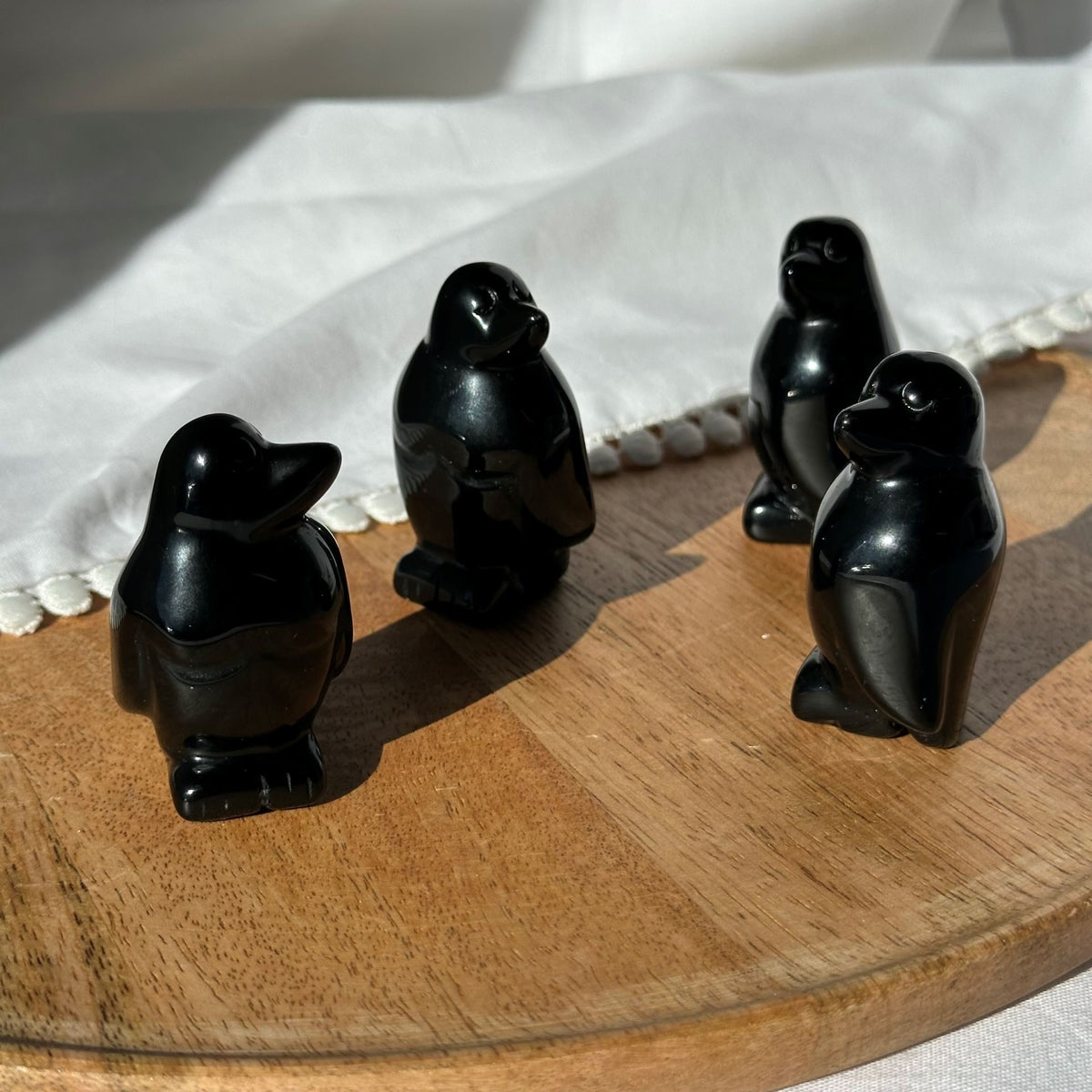 Fire små obsidian pingvin figurer står på en rund træplade, belyst af sollys.