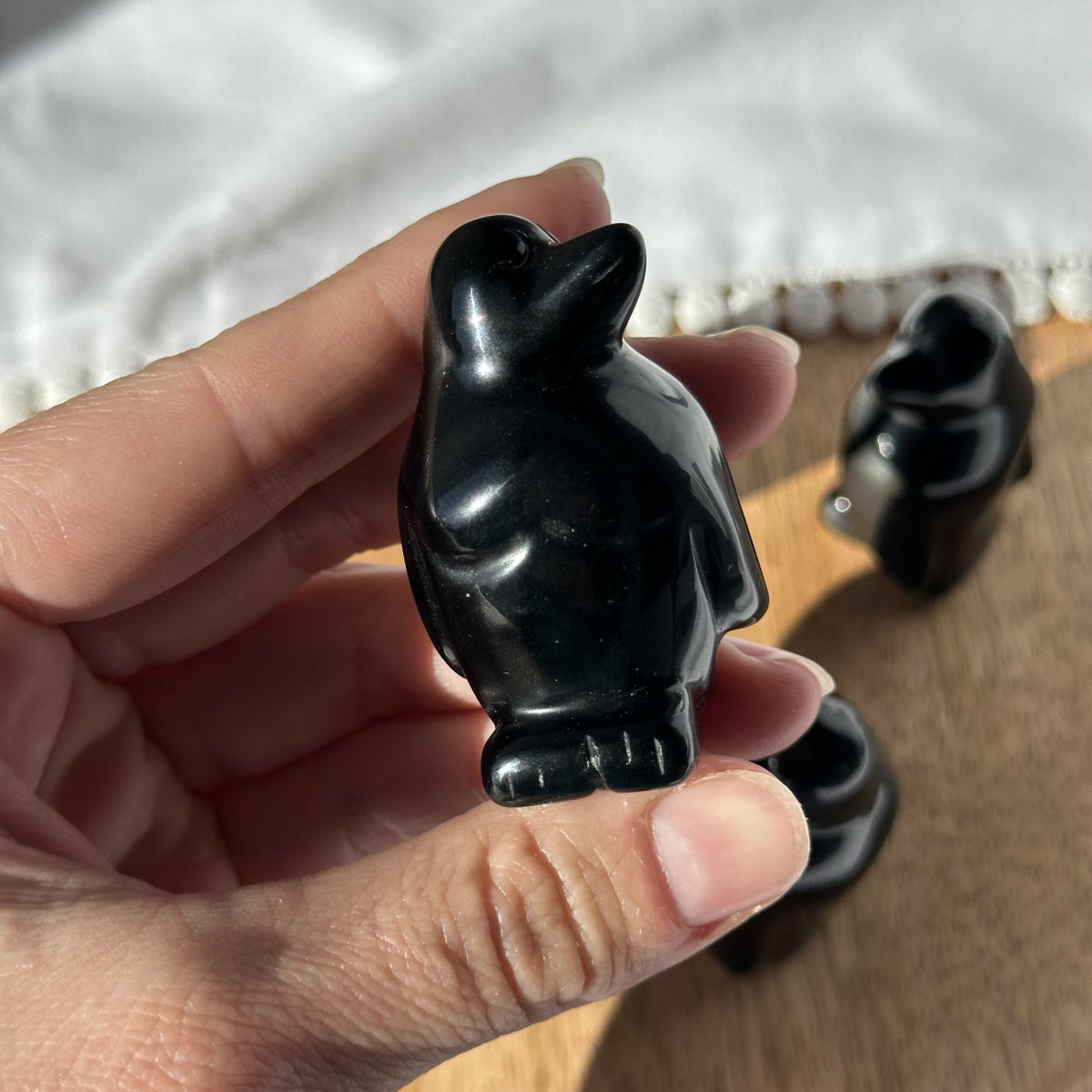 Hånd holder en lille skulptur af en obsidian pingvin, med bord og andre figurer i baggrunden.