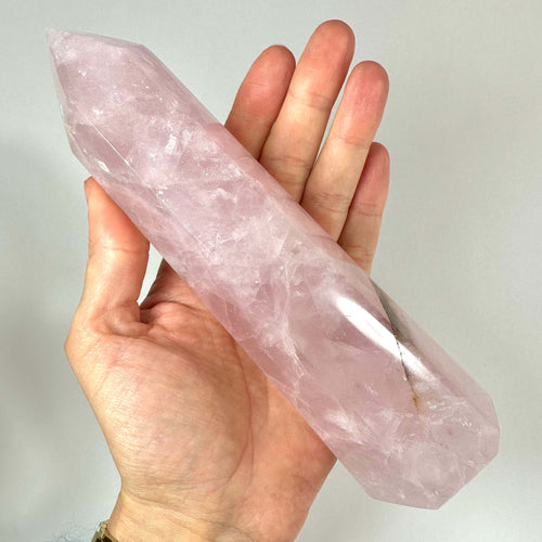 Et stort rosenkvarts tårn holdes i en hånd mod en neutral baggrund. Krystallen har en smuk, lyserød farve med naturlige inklusioner og en blank, slebet overflade. Tårnformen med spids top fremhæver krystallens æstetik og spirituelle formål. Rosenkvarts er kendt som kærlighedens krystal og forbindes med hjertchakraet, harmoni, medfølelse og følelsesmæssig heling.