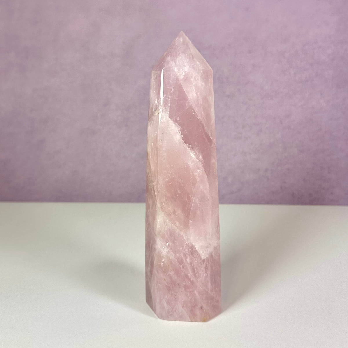 Et smukt rosenkvarts tårn står oprejst mod en lys lilla baggrund og en hvid overflade. Den lyserøde krystal er poleret med en glat, skinnende overflade og spidser til i en symmetrisk pyramideform. Kendt som kærlighedens sten symboliserer den selvomsorg, tilgivelse og følelsesmæssig heling. Et dekorativt og energisk redskab til meditation og harmoni.