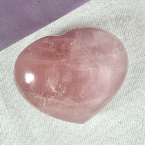 Rosenkvarts Hjerte XL Nr 2 er en smuk, stor lyserød krystal formet som et hjerte, placeret på en lys tekstilbaggrund med et lilla hjørne i toppen. Stenen symboliserer kærlighed, medfølelse og indre ro og er kendt for at styrke hjertechakraet. Perfekt til healing, meditation eller som dekorativt element med spirituel betydning.