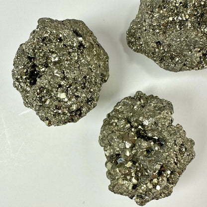 Tre skinnende pyrit klynger ligger på en hvid baggrund. De messinggule mineraler glitrer med deres karakteristiske metalliske glans og komplekse krystalstrukturer. Hver klynge består af utallige små krystaller, som fanger lyset og fremhæver deres guld-lignende udseende. Pyrit, også kaldet narreguld, er både et populært samlerobjekt og et spirituelt redskab med forbindelse til solar plexus chakraet.