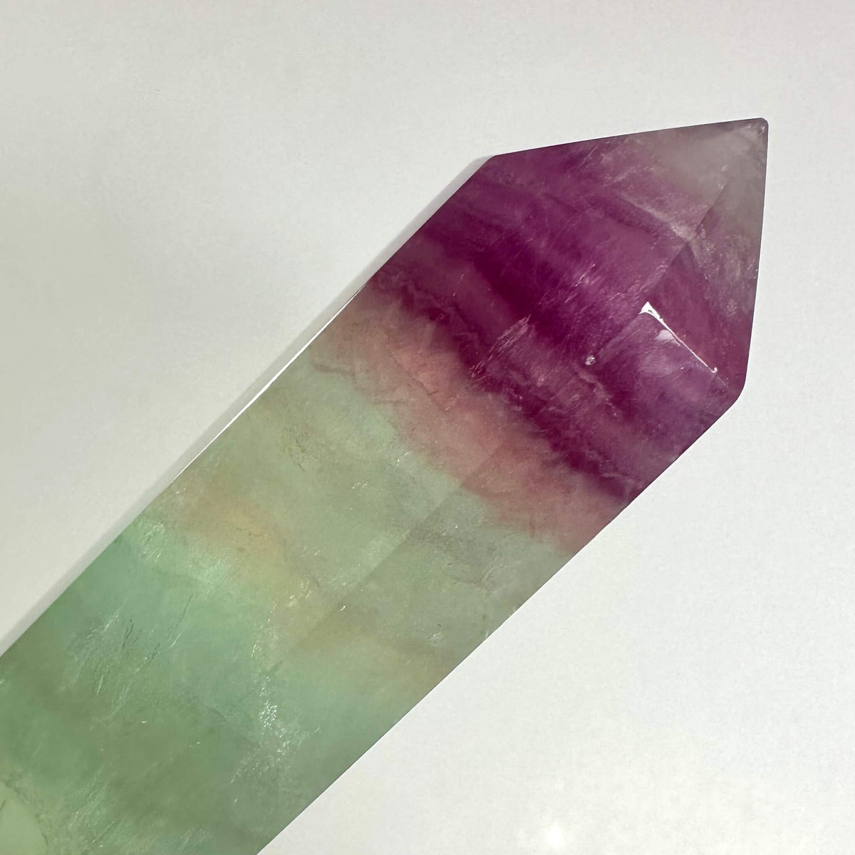 Et slankt, poleret Regnbue Fluorit tårn Nr 3 er vist mod en neutral baggrund. Krystallen fremviser smukke, naturlige lag i nuancer af lilla, grøn, rosa og let gul, som flyder harmonisk sammen. Dens spidse top og klare overflade fremhæver strukturens skønhed og energiske udstråling. Tårnet symboliserer balance og mental klarhed, ideel til meditation og spirituelt arbejde.