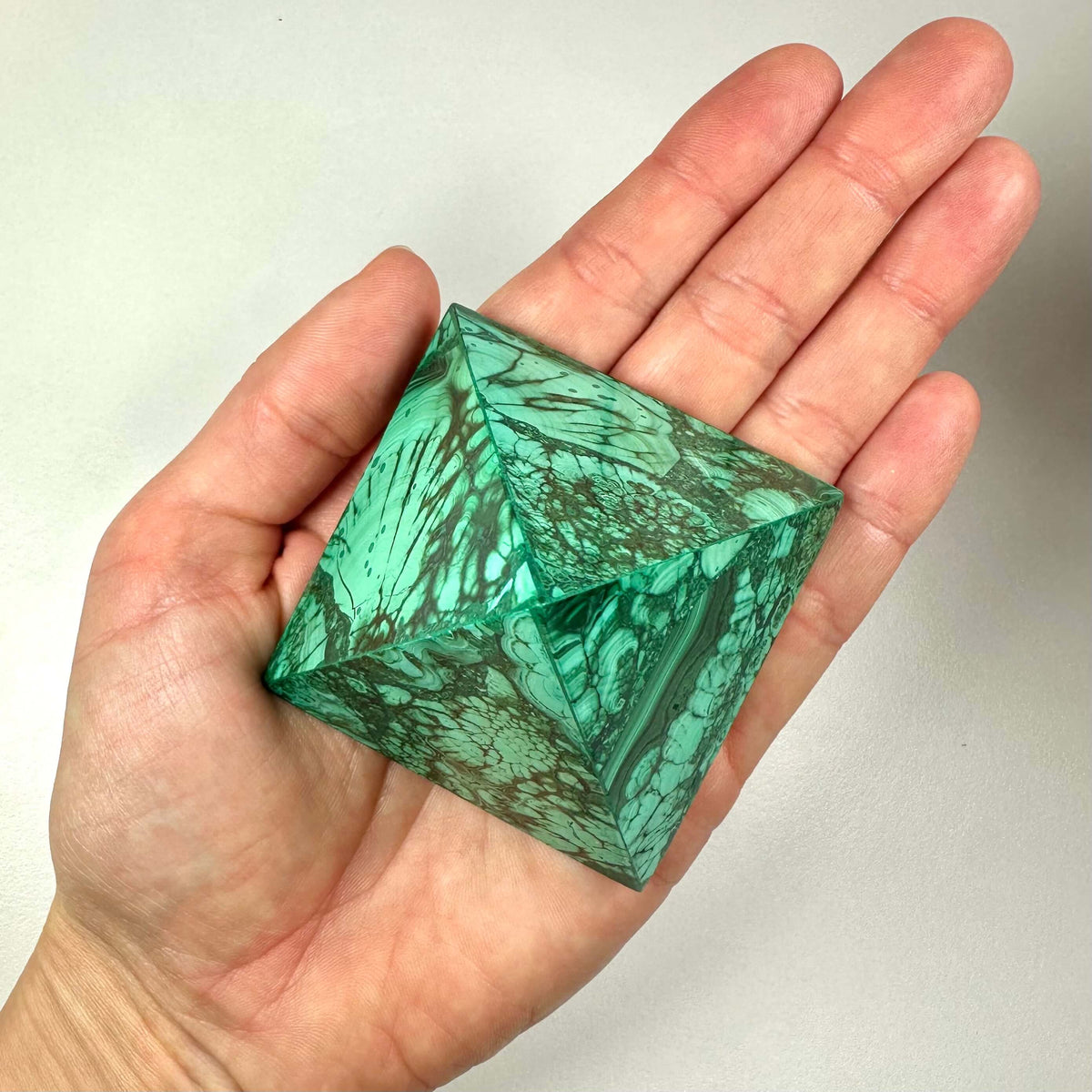 En hånd holder en poleret malakit pyramide med en levende grøn farve og karakteristiske mørkere bånd og mønstre. Malakit er kendt for sine spirituelle egenskaber, herunder beskyttelse, balance og følelsesmæssig healing, og forbindes især med hjertechakraet. Pyramideformen forstærker stenens energier, og denne version kombinerer æstetik med kraftfuld symbolik og spirituel anvendelse.