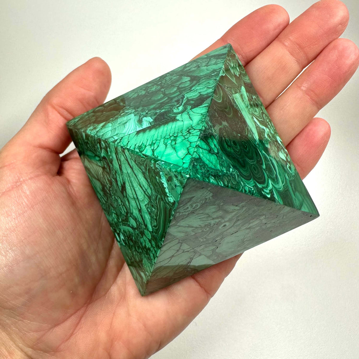 En hånd holder en smuk malakit pyramide, som fremstår med dybe grønne farver og karakteristiske båndmønstre. Den polerede overflade fremhæver stenens naturlige struktur og glans. Malakit pyramiden symboliserer transformation, healing og beskyttelse og er særligt brugbar i krystalhealing. Den fokuserede energi fra pyramideformen styrker følelsesmæssig balance og personlig udvikling.