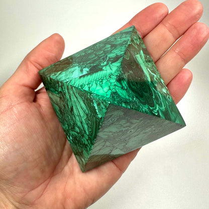 En hånd holder en smuk malakit pyramide, som fremstår med dybe grønne farver og karakteristiske båndmønstre. Den polerede overflade fremhæver stenens naturlige struktur og glans. Malakit pyramiden symboliserer transformation, healing og beskyttelse og er særligt brugbar i krystalhealing. Den fokuserede energi fra pyramideformen styrker følelsesmæssig balance og personlig udvikling.