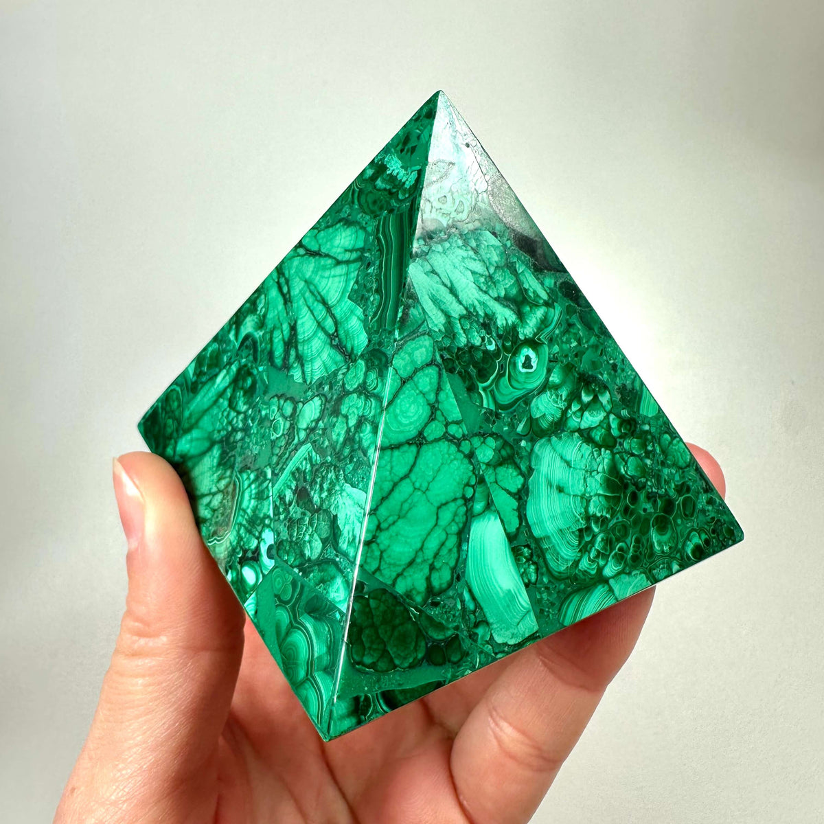 En hånd holder en smukt poleret malakit pyramide med dybgrønne nuancer og karakteristiske båndmønstre. Den glatte overflade reflekterer lyset og fremhæver stenens naturlige struktur. Malakit pyramiden symboliserer healing, beskyttelse og følelsesmæssig balance, og bruges ofte i krystalhealing. Den kraftfulde form kombineret med stenens energi gør den ideel til meditation og spirituelt arbejde.