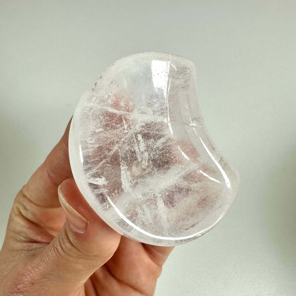 En hånd holder en smukt formet bjergkrystal smykkeskål, som er klar og gennemsigtig med fine naturlige linjer og indeslutninger. Skålen har en blød buet form og er lavet af rent siliciumdioxid, der symboliserer mental klarhed og åndelig renhed. Den anvendes ofte til opbevaring af smykker og som dekorativ genstand med balancerende energi i hjemmet.
