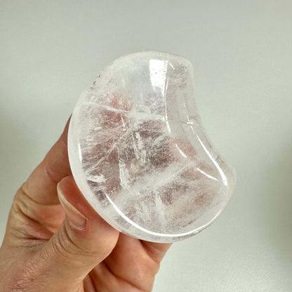 En hånd holder en smukt formet bjergkrystal smykkeskål, som er klar og gennemsigtig med fine naturlige linjer og indeslutninger. Skålen har en blød buet form og er lavet af rent siliciumdioxid, der symboliserer mental klarhed og åndelig renhed. Den anvendes ofte til opbevaring af smykker og som dekorativ genstand med balancerende energi i hjemmet.