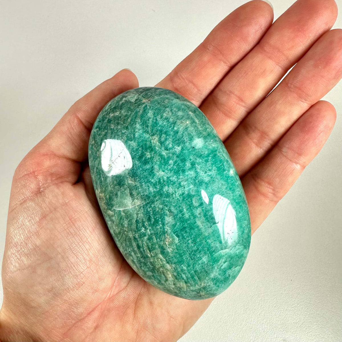 En hånd holder en smuk, poleret amazonit palmstone med en glat overflade og karakteristisk blågrøn farve. Stenen er oval og passer perfekt i håndfladen. Amazonit er kendt for sine beroligende og balancerende egenskaber, og bruges ofte i krystalhealing til at fremme kommunikation, reducere stress og åbne hjertechakraet. Billedet fremhæver stenens naturlige mønstre og glans.