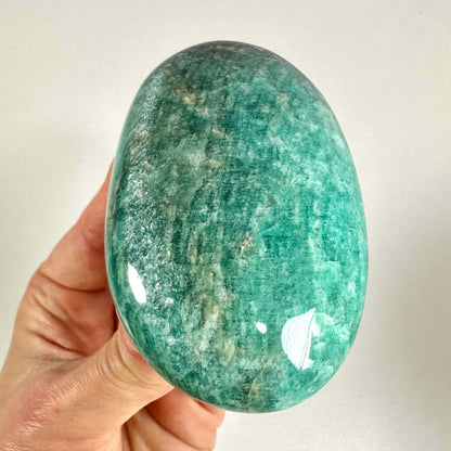En hånd holder en Amazonit palmstone med en glat, oval form og en iøjnefaldende blågrøn farve med naturlige mønstre. Stenen har en blank overflade, som reflekterer lyset smukt. Amazonit er kendt for sine beroligende og balancerende egenskaber, og bruges ofte i krystalhealing til at fremme ro, kommunikation og harmoni mellem hjerte og sind.