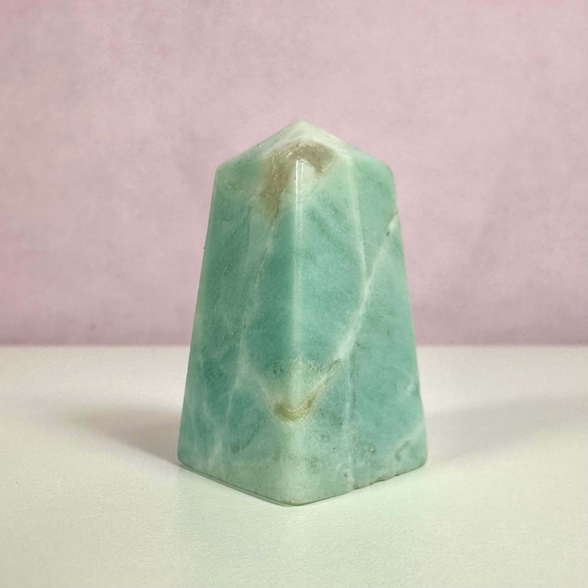Amazonit obelisk i blågrønne og turkise nuancer står mod en blød rosa baggrund. Den polerede krystal er formet som en obelisk og symboliserer balance, klarhed og kommunikation. Den bruges til meditation, chakra-healing og spirituelle praksisser. Farvespillet i stenen skyldes naturlige mineraler og gør den både æstetisk tiltalende og energisk kraftfuld. Perfekt som dekorativ og spirituel genstand.