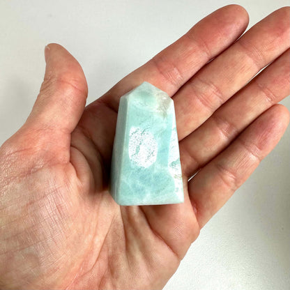 En hånd holder en glatpoleret amazonit obelisk med douce blågrønne nuancer og en spids top. Obelisken udstråler en rolig og beroligende energi og bruges ofte i meditation og chakra-healing. Den anses for at fremme kommunikation og balance, og dens kompakte størrelse gør den ideel som både spirituelt redskab og dekorativt element i hjemmet.