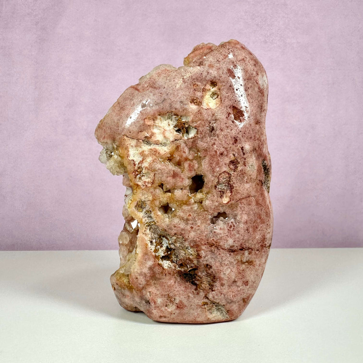 Pink ametyst, unika – en sjælden og smuk rå krystal med naturlige hulrum og nuancer i rosa og beige, stående mod en lyslilla baggrund. Denne unikke sten er kendt for sine healende egenskaber og forbindes især med hjertechakraet, hvor den fremmer kærlighed, balance og indre ro. Et dekorativt og spirituelt krystalstykke fra krystilriget.com.