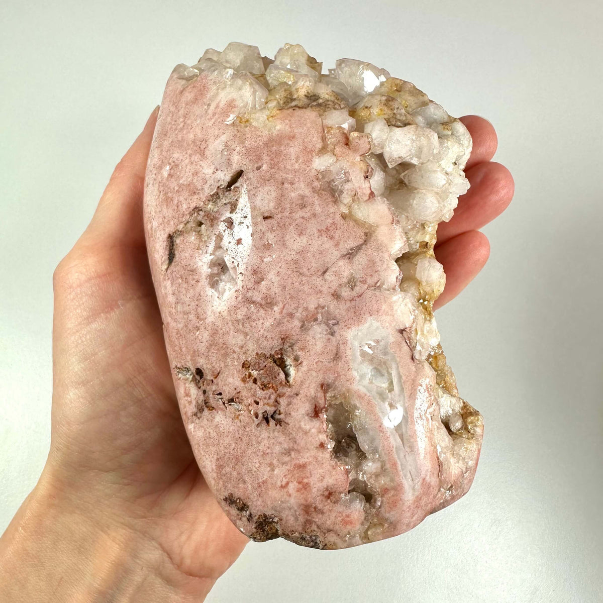 En hånd holder en stor, halvrå pink ametyst med en smuk, naturlig rosa farve og krystalliserede partier langs kanten. Stenen udviser en blanding af bløde nuancer og glitrende mineralaflejringer, hvilket understreger dens sjældenhed og æstetiske skønhed. Pink ametyst er kendt for sine healende egenskaber, især for hjertechakraet, og anvendes til følelsesmæssig balance og selvkærlighed.