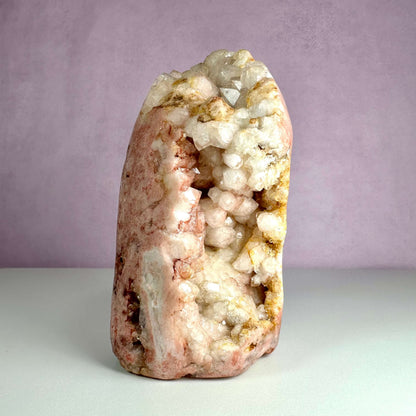 Unik og håndpoleret pink ametystkrystal stående lodret mod en lyserød baggrund. Krystallen har en naturlig, rå struktur med glitrende druzy-lommer og nuancer af rosa, hvid og gyldenbrun. Den fremstår kraftfuld og æstetisk, ideel til hjertehealing og spirituelle praksisser. En sjælden skønhed fra Patagonien, som kombinerer visuel elegance med følelsesmæssig ro.