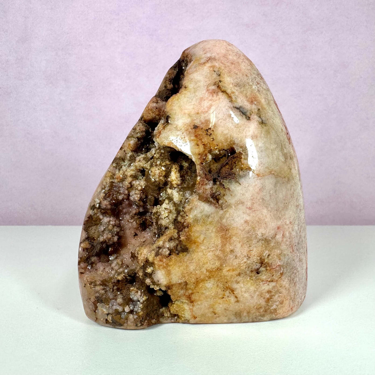 En unik, fritstående pink ametystkrystal med glatpolerede overflader og naturlige geodeformationer. Stenen har varme nuancer af rosa, beige og brun med synlige krystalformationer i en åbning. Baggrunden er enkel og lyserød, hvilket fremhæver stenens naturlige skønhed. Krystallen er kendt for sine beroligende egenskaber og anvendes til emotionel balance, meditation og hjertechakraets aktivering.