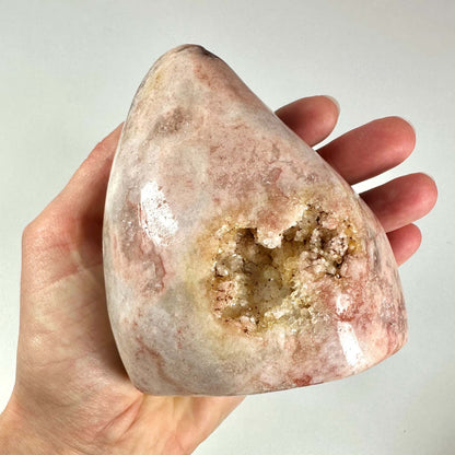 En hånd holder en unik, poleret pink ametyst med en naturlig geodeåbning fyldt med glitrende krystaller. Stenen har bløde rosa og beige nuancer med svage årer fra hematit-inklusionsdannelse, som giver et harmonisk, beroligende udtryk. Pink ametyst er kendt for sine healende egenskaber som fremmer følelsesmæssig balance, selvkærlighed og spirituel klarhed, og er et eftertragtet krystalobjekt.