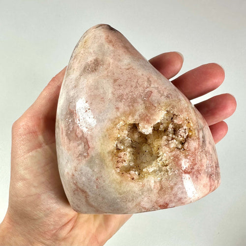 En hånd holder en unik, poleret pink ametyst med en naturlig geodeåbning fyldt med glitrende krystaller. Stenen har bløde rosa og beige nuancer med svage årer fra hematit-inklusionsdannelse, som giver et harmonisk, beroligende udtryk. Pink ametyst er kendt for sine healende egenskaber som fremmer følelsesmæssig balance, selvkærlighed og spirituel klarhed, og er et eftertragtet krystalobjekt.