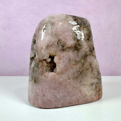 En unik, poleret Pink Ametyst-sten står mod en lyserød baggrund. Stenen har en silkeblød overflade i sarte nuancer af rosa og grå, med naturlige hulrum og krystaller synlige i midten. Denne sten symboliserer kærlighed, heling og ro, og er ideel til meditation og dekoration. Et kraftfuldt stykke, der arbejder med hjertechakraet og beskytter mod negativ energi.