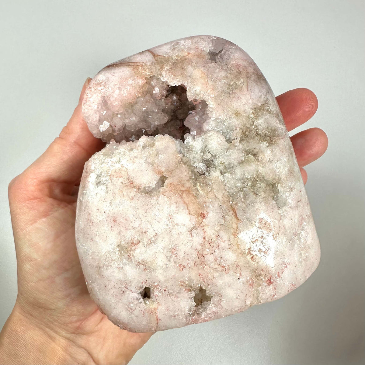 En hånd holder en stor, poleret Pink Ametyst sten med en åben krystalformation i midten. Den unikke sten har en delikat rosa nuance med indslag af hvidt og let brunlige mineralårer. Overfladen er delvist dækket af glitrende krystaller, som fremhæver stenens naturlige skønhed. Stenen symboliserer kærlighed, heling og spirituel ro og arbejder især med hjertechakraet.