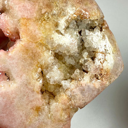 Nærbillede af en sjælden Pink Ametyst, unika, med en blanding af rosa, fersken og gyldne nuancer samt en krystallinsk druzy-overflade i midten. Krystallen er naturligt formet og fremstår rå og ubehandlet med funklende kvartskrystaller. Pink ametyst er kendt for sine healende egenskaber og forbindes med hjertchakraet, samt følelsesmæssig balance og spirituel vækst.