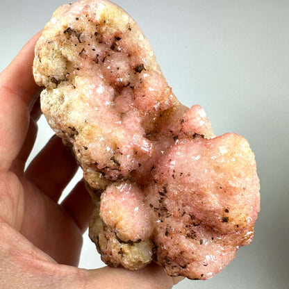 En smuk og sjælden Pink Ametyst-krystal holdes i hånden mod en neutral baggrund. Krystallen udviser strålende rosa og ferskenfarver med glitrende overflader og naturlige indeslutninger af hæmatit. Dens unikke geologiske formation fremhæver dens naturlige kunstneriske udtryk. Pink Ametyst er kendt for sine healende egenskaber, der fremmer kærlighed, følelsesmæssig balance og spirituel vækst.
