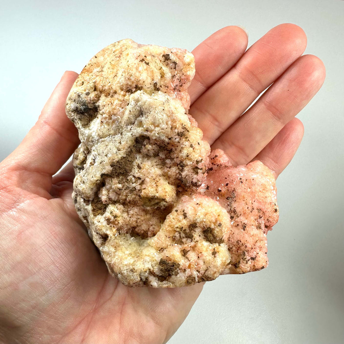 En hånd holder en stor, ujævn pink ametyst krystal med naturlige formationer i nuancer af rosa, fersken og beige. Krystallen har en rå overflade med glitrende druzy-strukturer, og mørkere indeslutninger ses spredt over det hele. Pink ametyst er en sjælden variant af kvarts, kendt for sine healende egenskaber og evne til at fremme kærlighed og følelsesmæssig balance.