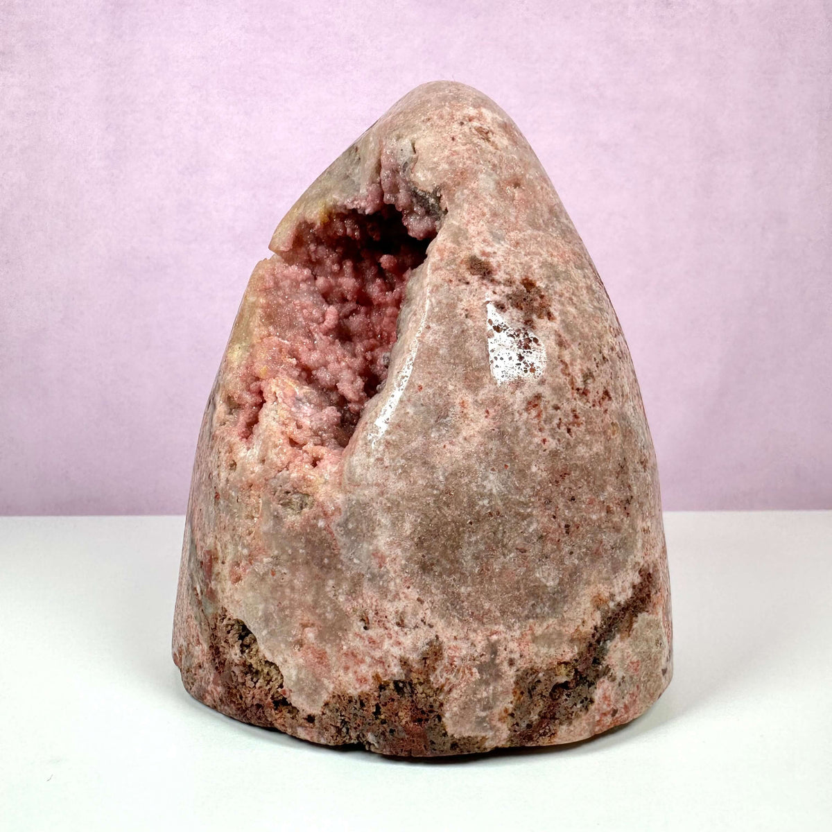 Unik Pink Ametyst geode stående mod en lys rosa baggrund. Krystallen har en poleret ydre overflade med naturlige rødbrune og lyserøde nuancer, mens indersiden afslører glitrende krystaller i sarte pink toner. Den sjældne sten repræsenterer kærlighed, medfølelse og følelsesmæssig healing, og er især værdsat for sin skønhed og spirituelle energi. Perfekt til meditation og dekoration.