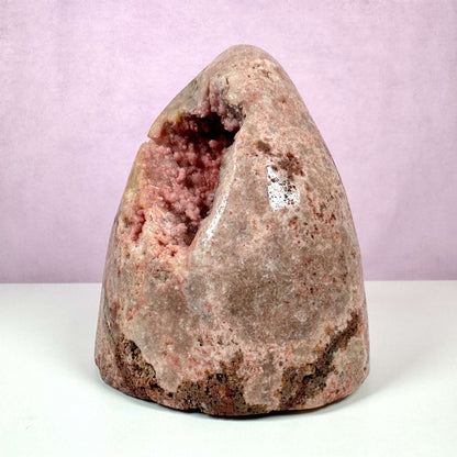 Unik Pink Ametyst geode stående mod en lys rosa baggrund. Krystallen har en poleret ydre overflade med naturlige rødbrune og lyserøde nuancer, mens indersiden afslører glitrende krystaller i sarte pink toner. Den sjældne sten repræsenterer kærlighed, medfølelse og følelsesmæssig healing, og er især værdsat for sin skønhed og spirituelle energi. Perfekt til meditation og dekoration.