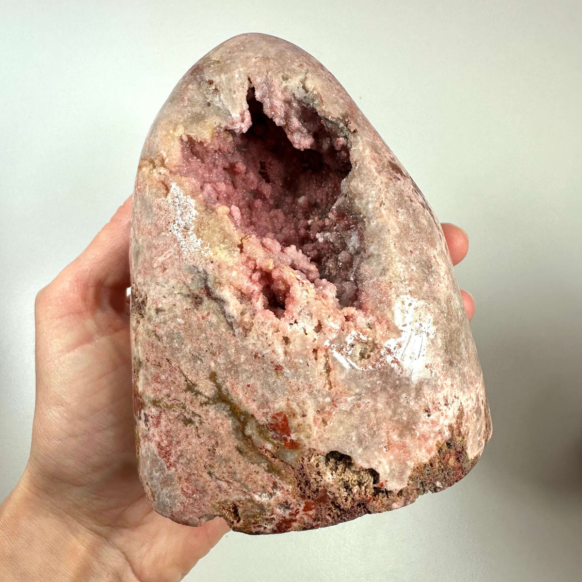 En hånd holder en stor, poleret pink ametyst med en naturlig hulning fyldt med glitrende krystaller i sarte rosa nuancer. Stenen udviser flotte farvevariationer fra fersken til dyb pink og indeholder naturlige mønstre og strukturer, typiske for ægte ametyst med hæmatitindhold. Dette unika-stykke symboliserer kærlighed, ro og spirituel dybde, og er ideelt til meditation og følelsesmæssig healing.