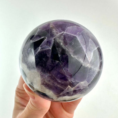 En hånd holder en Chevron Ametyst kugle, som fremviser smukke lagdelte mønstre i violette og hvide nuancer. Krystallen har en blank, poleret overflade, der reflekterer lyset og fremhæver dens unikke struktur. Kuglen symboliserer spirituel skønhed og bruges ofte i meditation og healing for at skabe ro og klarhed samt beskytte mod negativ energi.