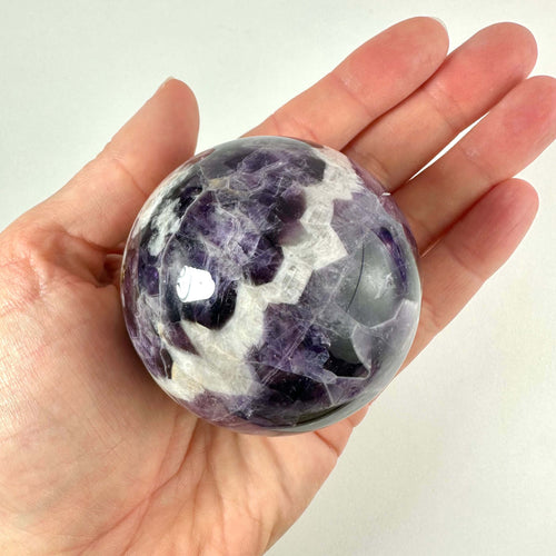 En hånd holder en smuk, poleret Chevron Ametyst kugle med karakteristiske violette og hvide V-formede bånd. Kuglen fremstår glansfuld og unik med sit naturlige mønster og farvespil. Den spirituelle sten siges at bringe ro og klarhed, samt beskytte mod negativ energi. Baggrunden er neutral, hvilket fremhæver kuglens detaljer og æstetiske kvaliteter.