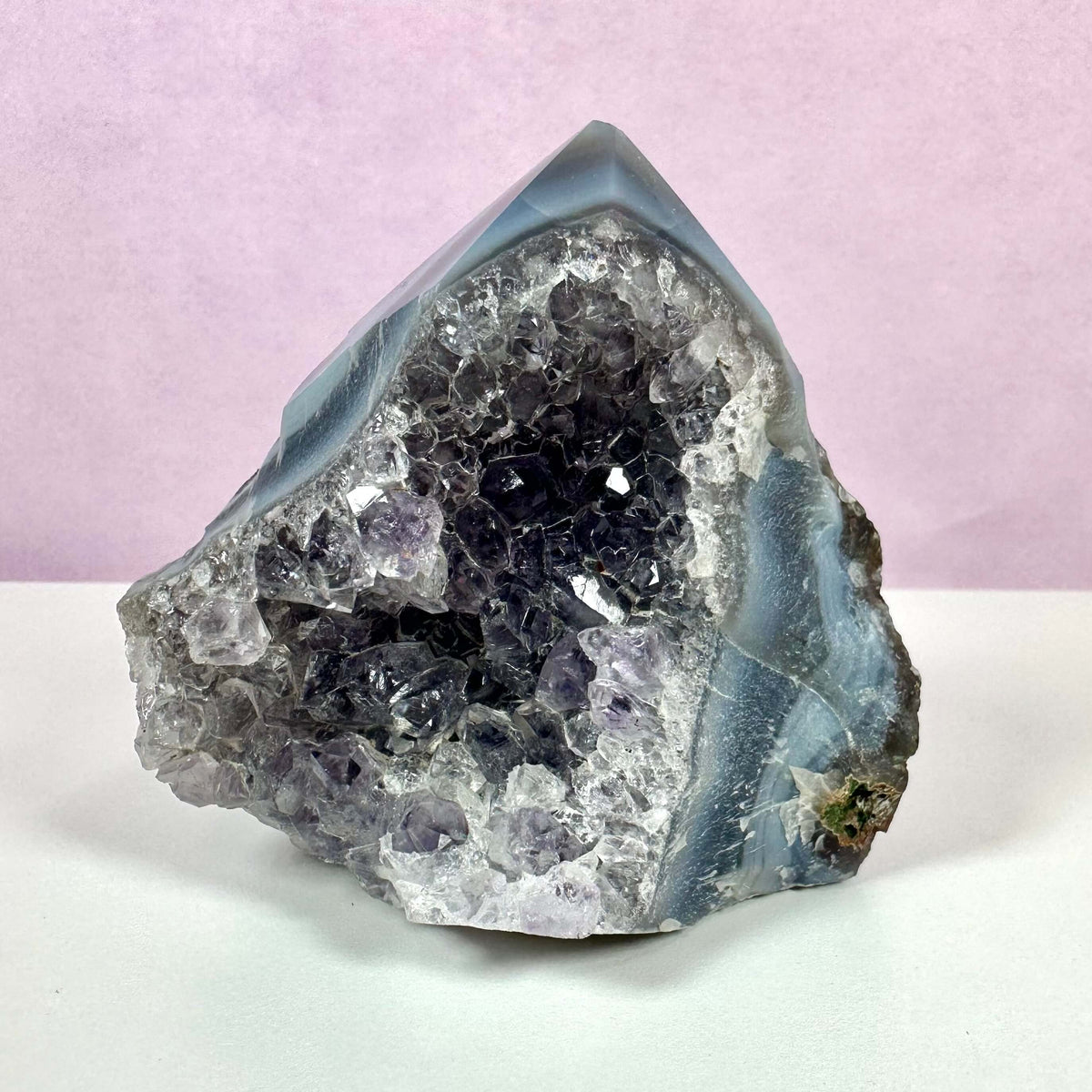 Drusy Ametyst spids med en krystalliseret, glitrende overflade i dybe lilla og klare nuancer. Krystallen står på en hvid overflade med en lyslilla baggrund. Dens naturlige formation og spidse top gør den ideel til meditation og energiarbejde. Den kombinerer æstetisk skønhed med spirituelle egenskaber og anvendes ofte til at fremme ro, mental klarhed og spirituel bevidsthed.