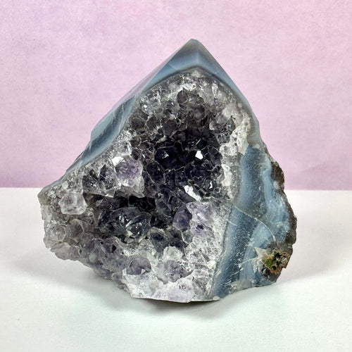 Drusy Ametyst spids med en krystalliseret, glitrende overflade i dybe lilla og klare nuancer. Krystallen står på en hvid overflade med en lyslilla baggrund. Dens naturlige formation og spidse top gør den ideel til meditation og energiarbejde. Den kombinerer æstetisk skønhed med spirituelle egenskaber og anvendes ofte til at fremme ro, mental klarhed og spirituel bevidsthed.