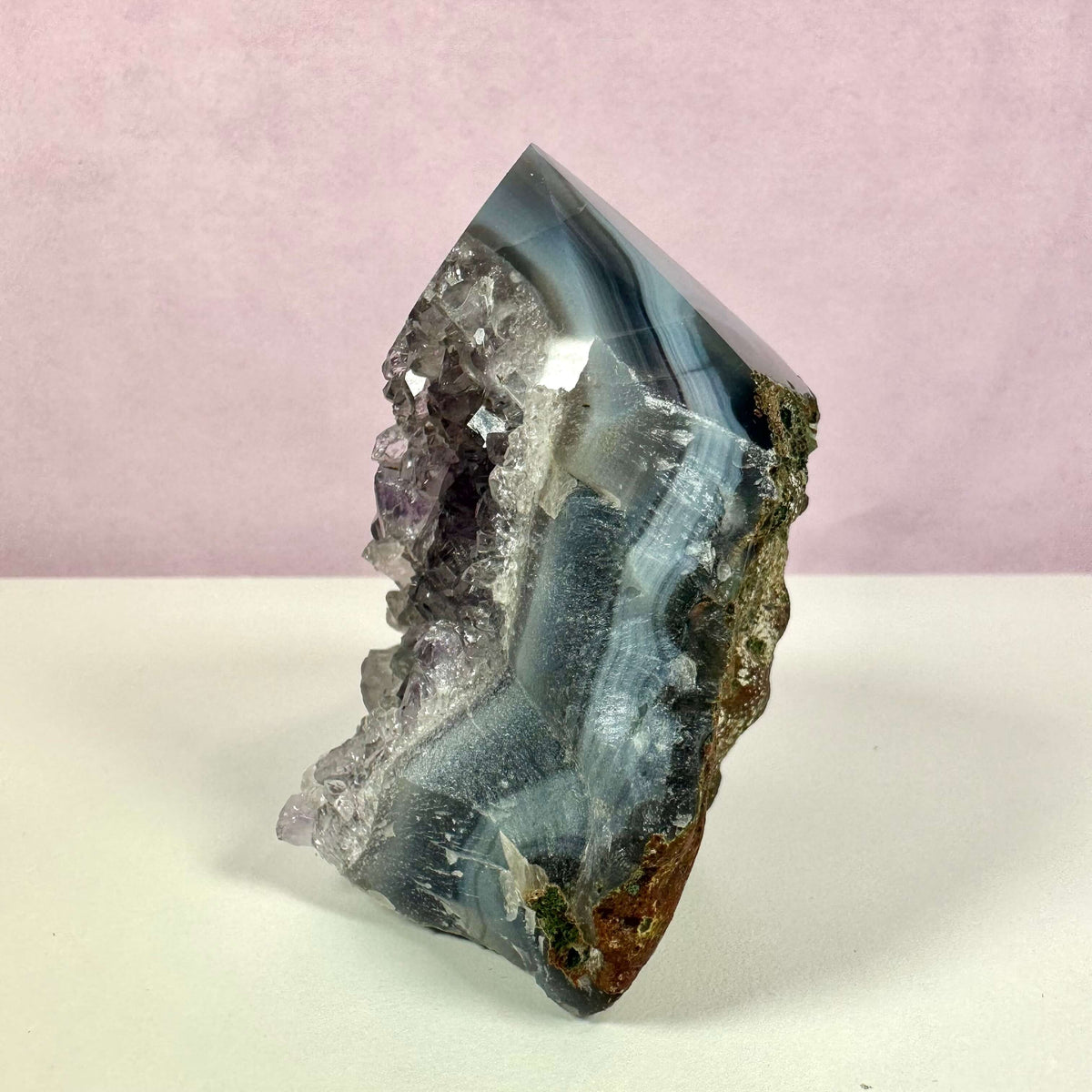 Drusy Ametyst spids vist mod en blød lyserød baggrund. Den polerede spids fremhæver de dybe lilla toner i ametysten, mens den glitrende drusy-overflade med små krystaller giver et funklende udtryk. Krystallen har naturlige lag i grå, blå og hvide nuancer samt rå kanter i brun og grøn. Ideel til meditation, spirituel praksis og dekorativ indretning.