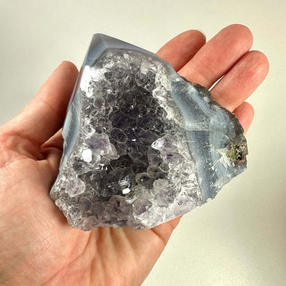 En hånd holder en Drusy Ametyst spids, der udstråler en smuk lilla glans med en glitrende krystaloverflade. Spidsen har et naturligt, råt udseende og fremviser tydeligt de mange små ametystkrystaller, der dækker overfladen. Krystallen kombinerer indre klarhed med spirituel energi og bruges ofte i meditation og healing. Baggrunden er lys og neutral.