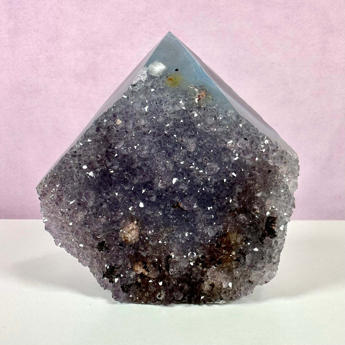 Drusy ametyst spids vist mod en lyserød baggrund. Krystallen har en poleret spids og en glitrende, ru drusy-overflade med lilla og violette nuancer. De små kvarts-krystaller fanger lyset smukt og giver stenen et levende, funklende udtryk. Perfekt til meditation og spirituel praksis, da den kombinerer æstetisk skønhed med beroligende energi og åndelig fordybelse.